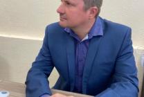 тифлокомментарий. На фотографии изображен председатель Петрозаводской МО ВОС. Степан Валерьевич Дьяконов сидит за столом. На нём надета синяя рубашка с узором и синий пиджак. Мужчина смотрит влево. Его руки сложены на столе.